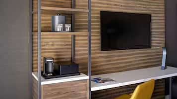 A modern hotel room with a wooden wall panel, a flat-screen TV mounted on the wall, a white desk with a magazine, a coffee maker, and a yellow chair.