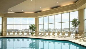 Una gran piscina interior con numerosas tumbonas y amplios ventanales con vistas a edificios.