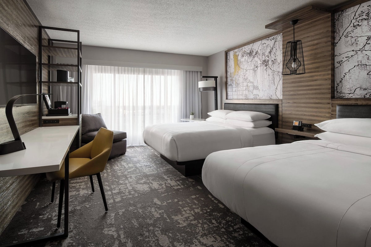 A hotel room with a desk, two beds, a chair, and a large window with curtains.