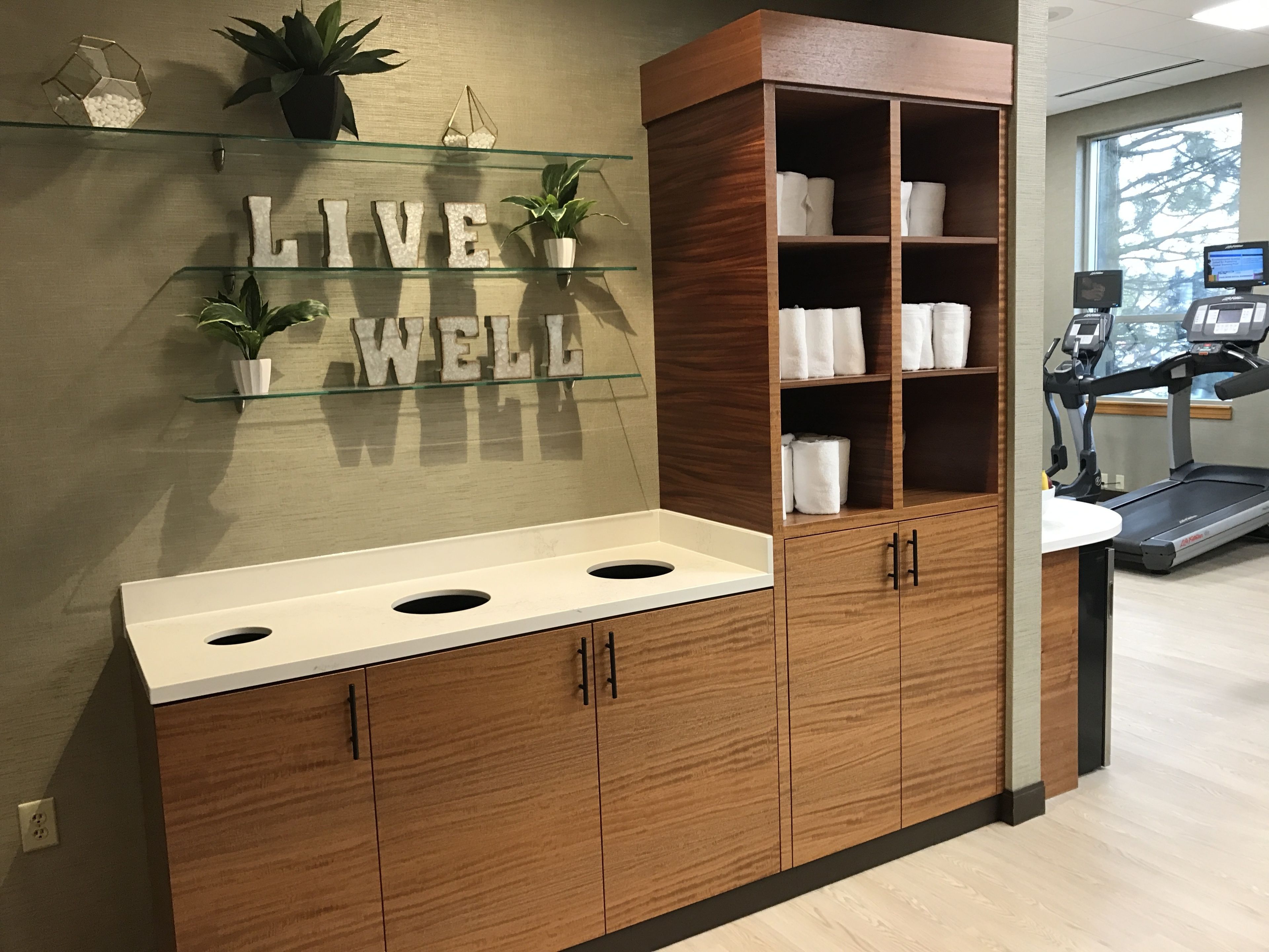 A bathroom with a wooden vanity, a glass shelf with plants, and a motivational sign saying "LIVE WELL".