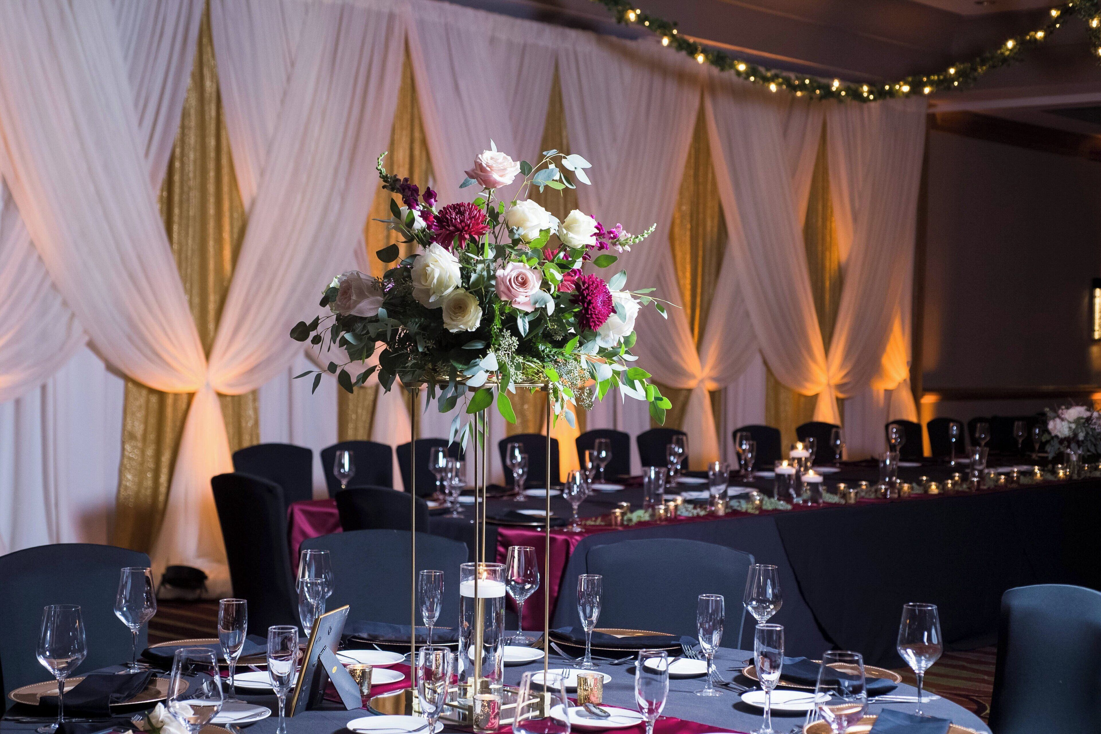 A banquet hall with tables set for a formal event, featuring a central floral arrangement and draped curtains in the background.