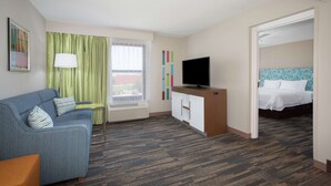 Habitación de hotel con sofá azul, televisor sobre un mueble de madera, cama con ropa blanca y ventana con cortinas verdes.