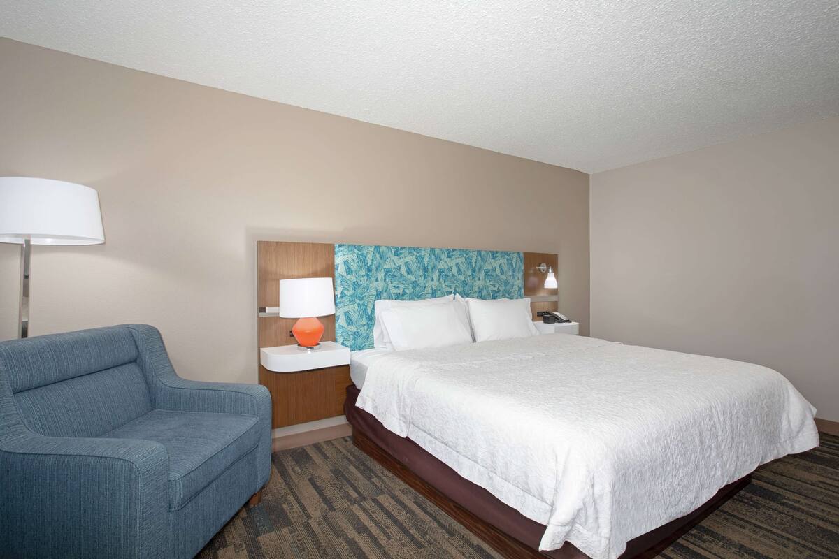 A hotel room with a bed, a blue armchair, a nightstand with a lamp, and a headboard with a blue pattern.