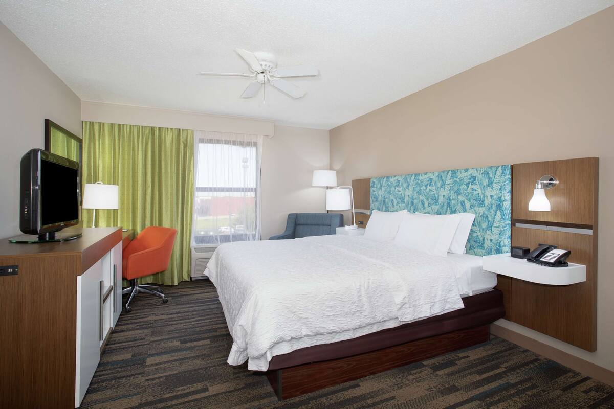 A hotel room with a large bed, a desk with a television, a chair, a desk lamp, a phone, and a ceiling fan.