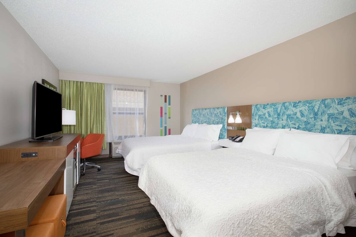 A hotel room with two beds, a desk, a television, and a window with curtains.