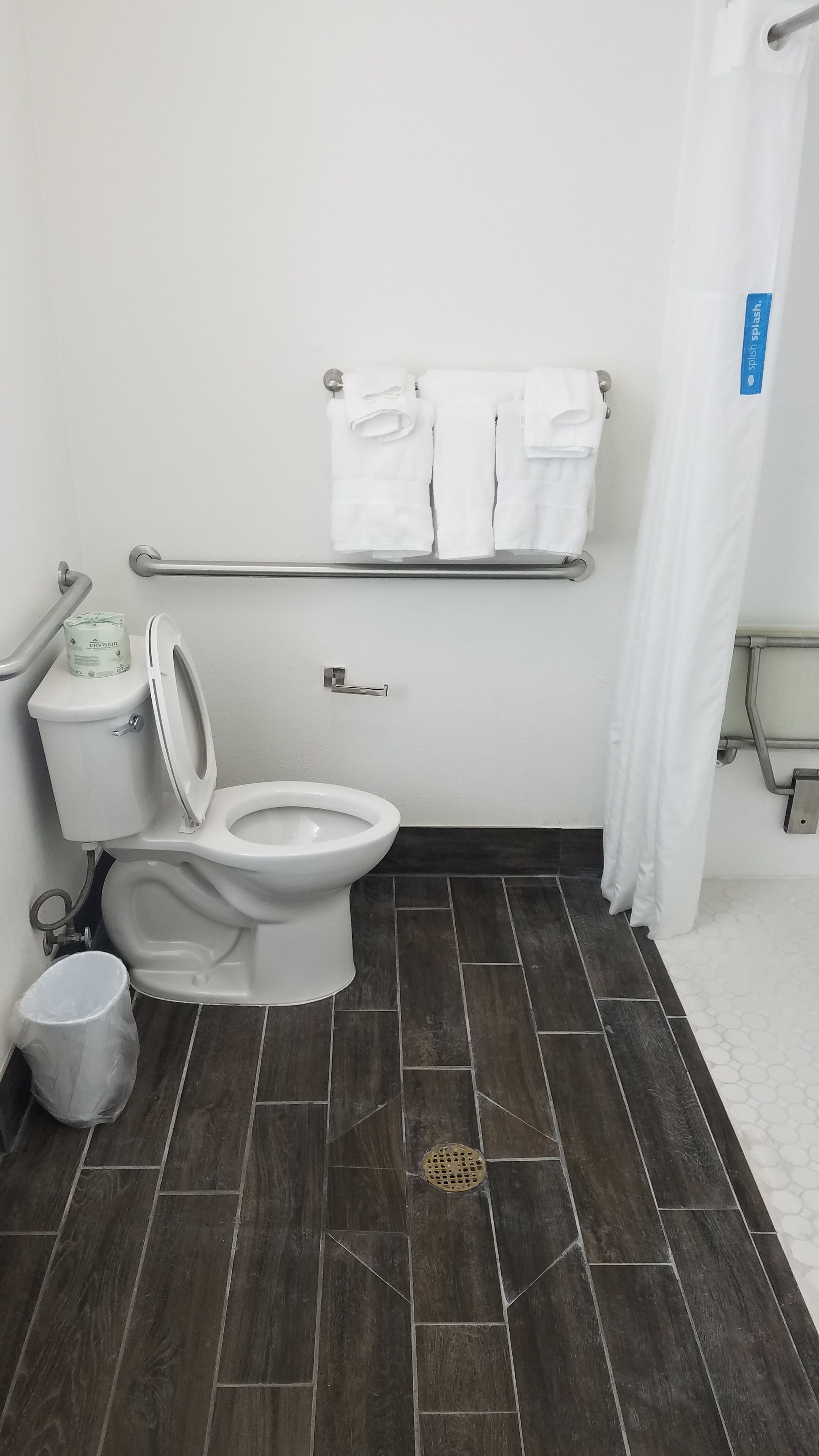 A bathroom with a toilet, a handrail, a towel rack with towels, a trash can, and a shower curtain.