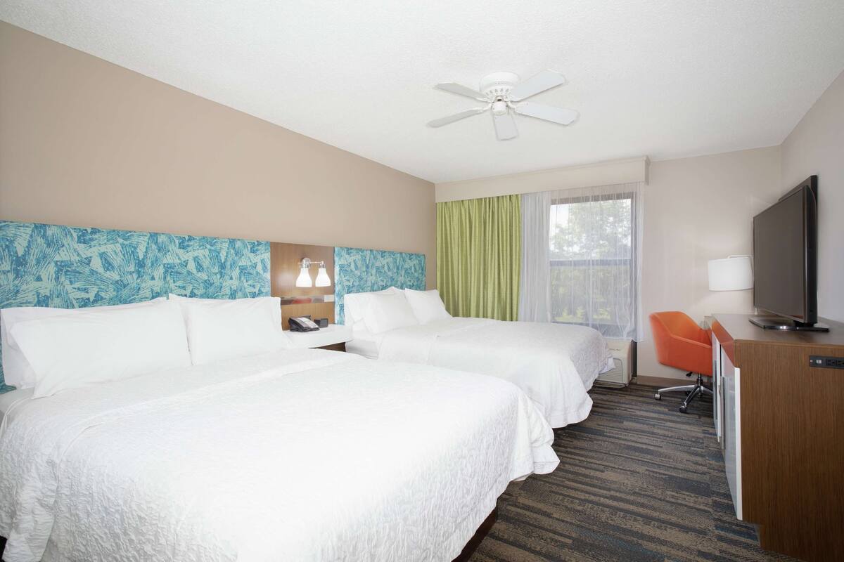 A hotel room with two beds, a desk with a television, a desk lamp, and a ceiling fan.