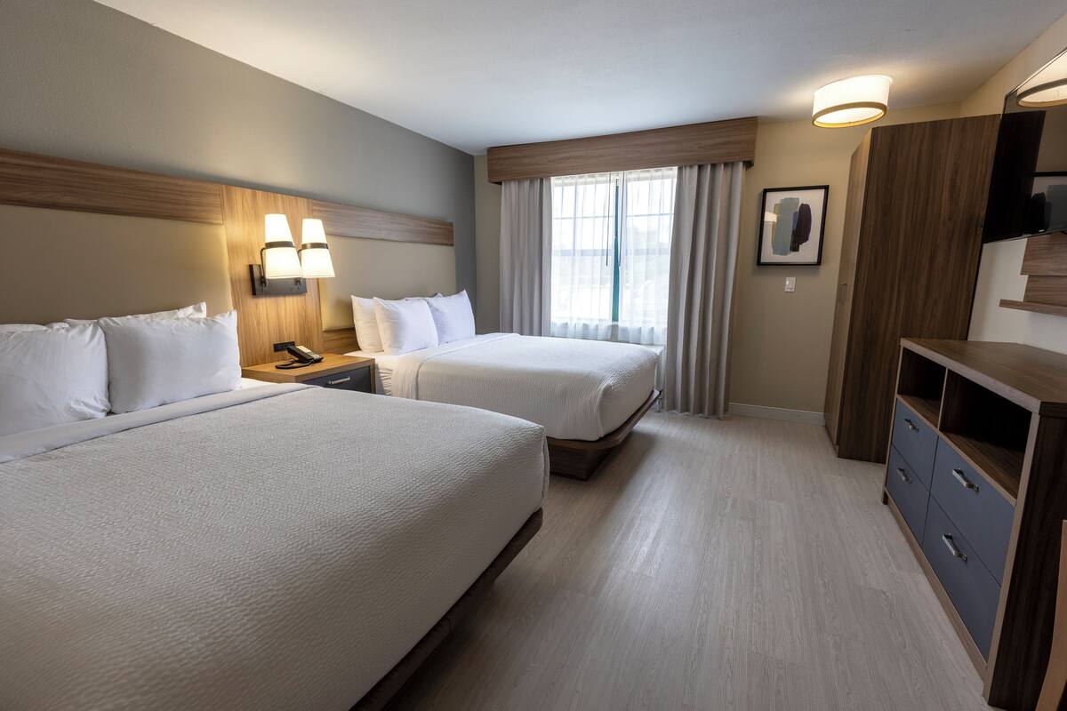 Ein Hotelzimmer mit zwei Betten, einem hölzernen Kopfteil, einem Nachttisch, einem Schreibtisch, einem Fernseher und einem großen Fenster mit Vorhängen.