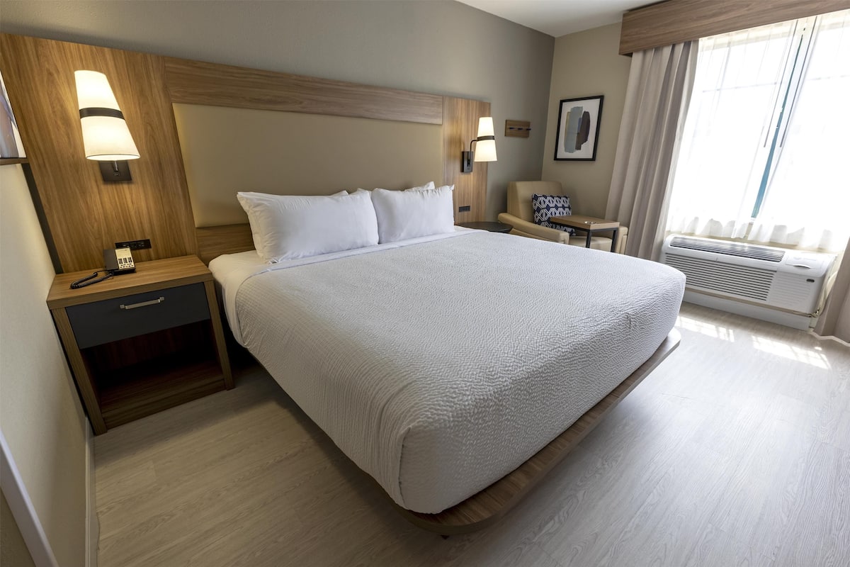 Ein Hotelzimmer mit einem großen Bett, Nachttischen, einem Sessel, einem Schreibtisch und einem fenster mit Vorhängen.