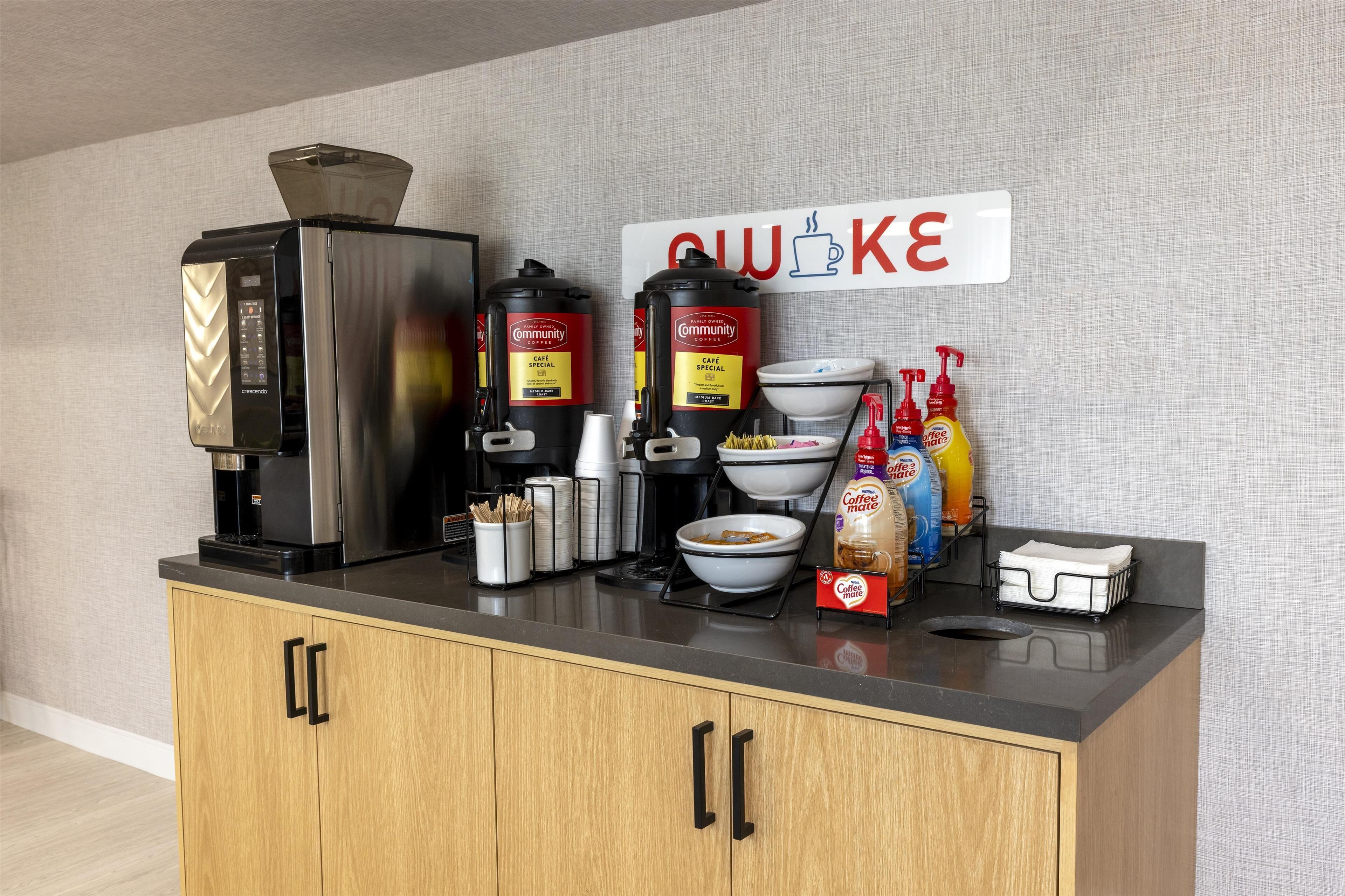 A kitchenette with a coffee maker, two coffee dispensers, a bowl, a coffee mug, and a sign that says "CUCKE".