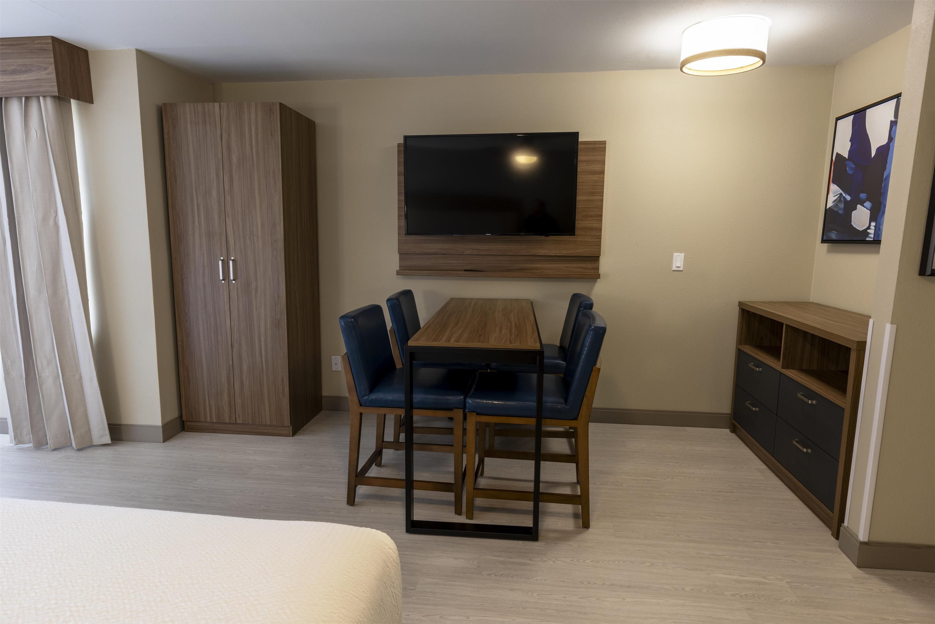 Hotelkamer met een houten eettafel, blauwe stoelen, een aan de muur bevestigde televisie, een garderobe en een klein kastje.