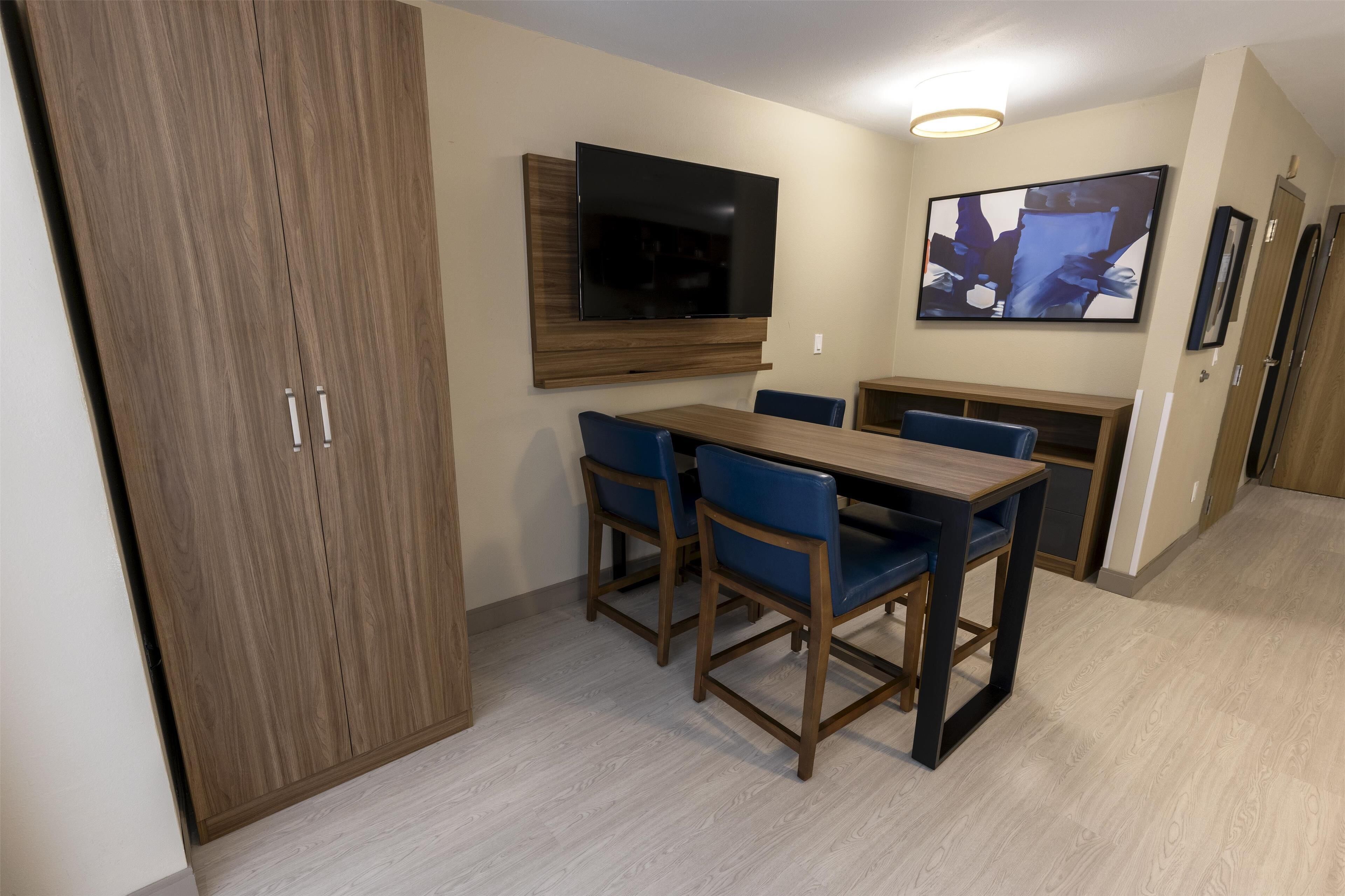 Un comedor con una mesa de madera y sillas azules, un televisor de pantalla plana montado en la pared y un cuadro enmarcado sobre el televisor.