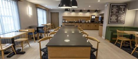 Un comedor moderno con una mesa comunal larga, varias sillas y una zona de cocina con cafetera.