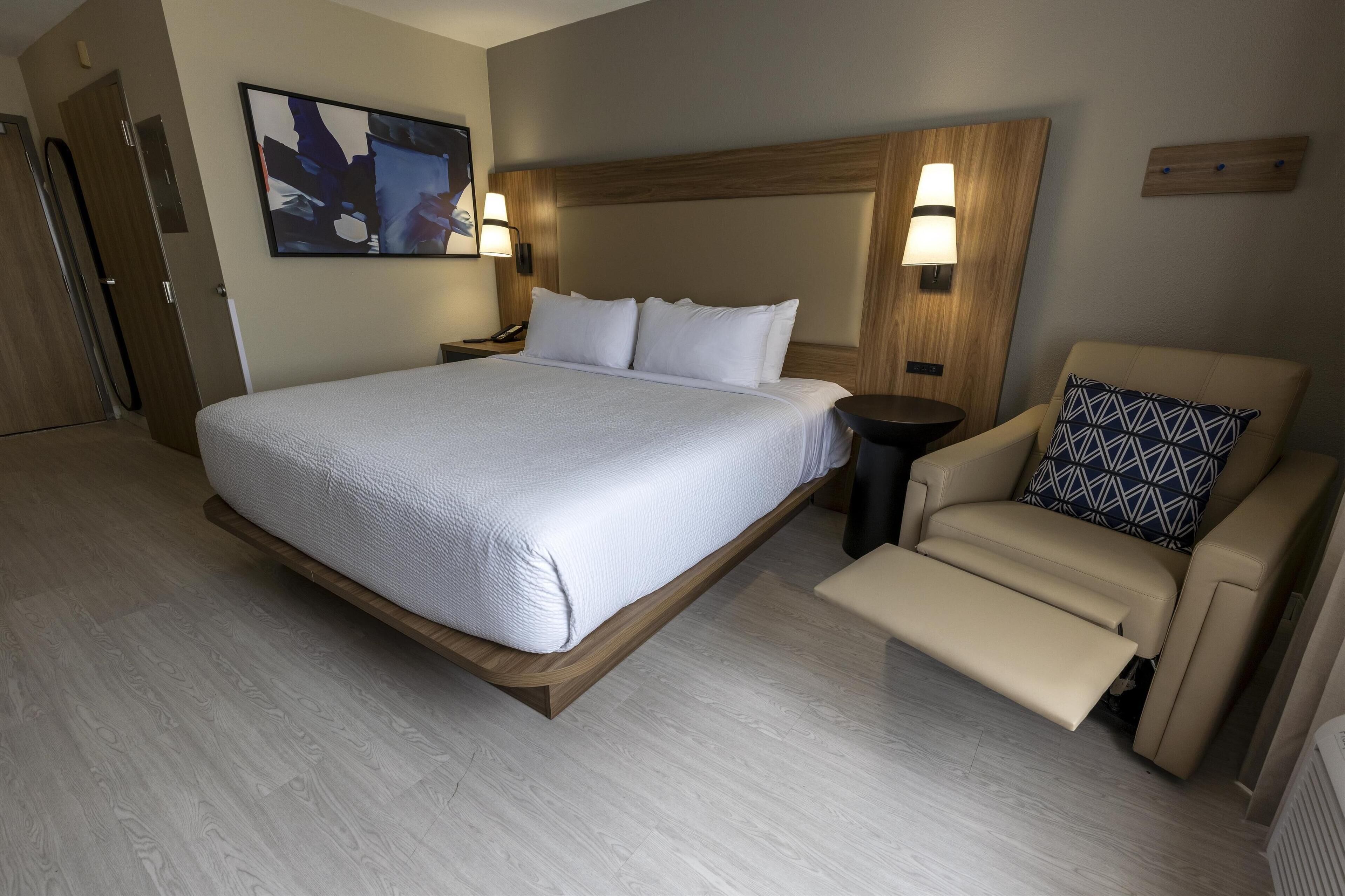 A hotel room with a large bed, a bedside table, a chair with a patterned pillow, and a wall-mounted light fixture.
