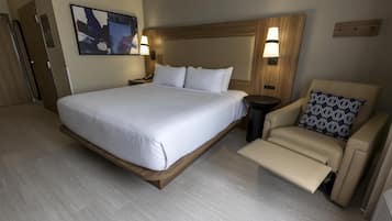 A hotel room with a large bed, a bedside table, a chair with a patterned pillow, and a wall-mounted light fixture.