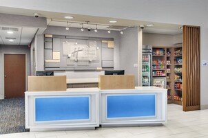 Un mostrador de recepción de hotel con diseño moderno, un refrigerador lleno de bebidas y un bar de snacks con distintos productos.