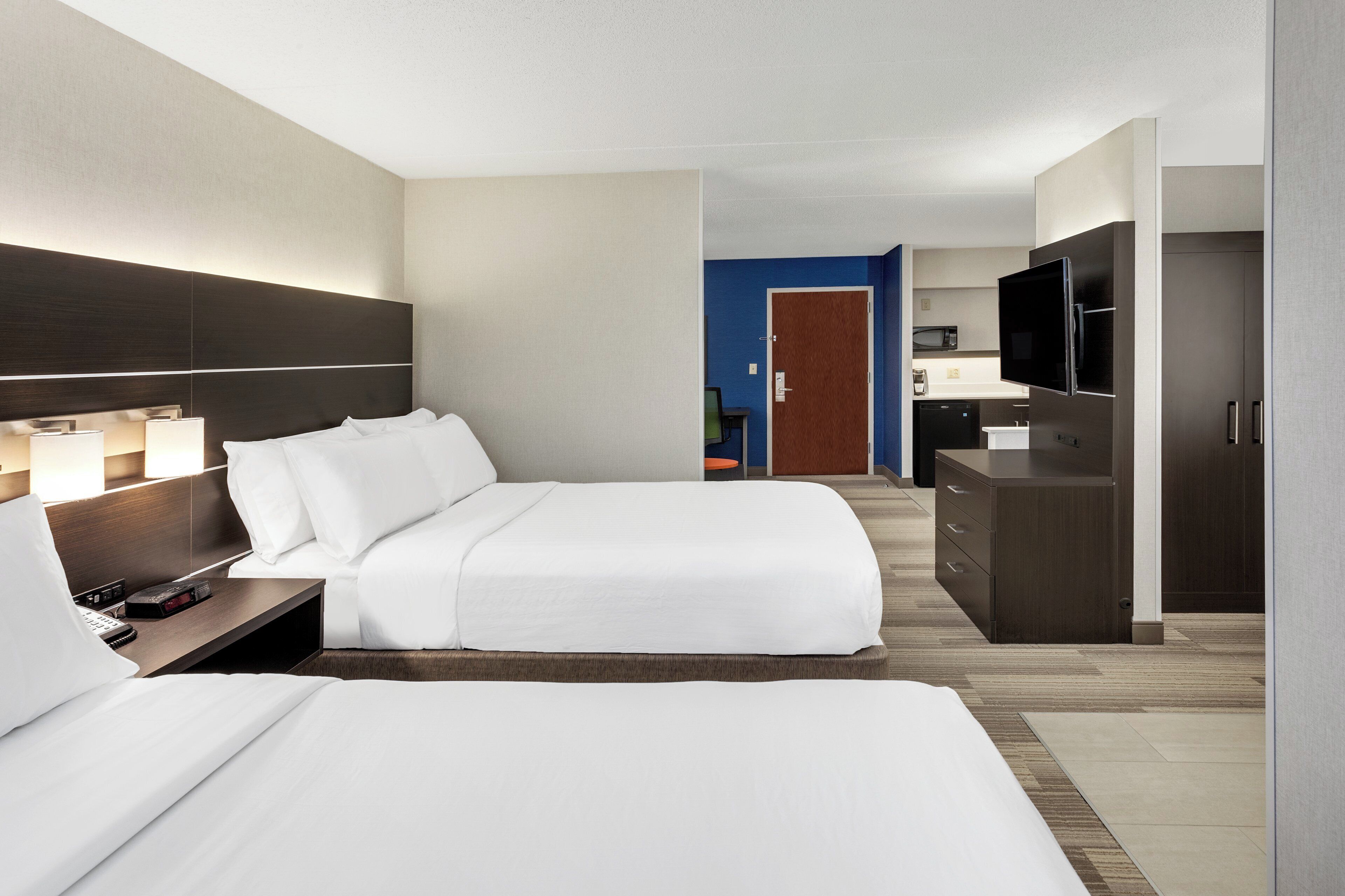A hotel room with two beds, a desk with a lamp, a TV, and a kitchenette.