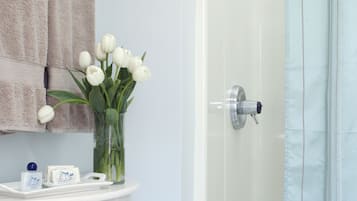 Une salle de bain avec un lavabo blanc, un miroir, un porte-serviettes avec une serviette, un vase en verre avec des fleurs et un espace douche doté d’une porte en verre.