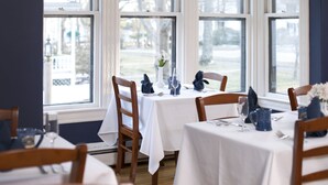 A dining area with white tablecloths, wooden chairs, and a view of a park through large windows. - Brewster House Bed & Breakfast (Freeport)