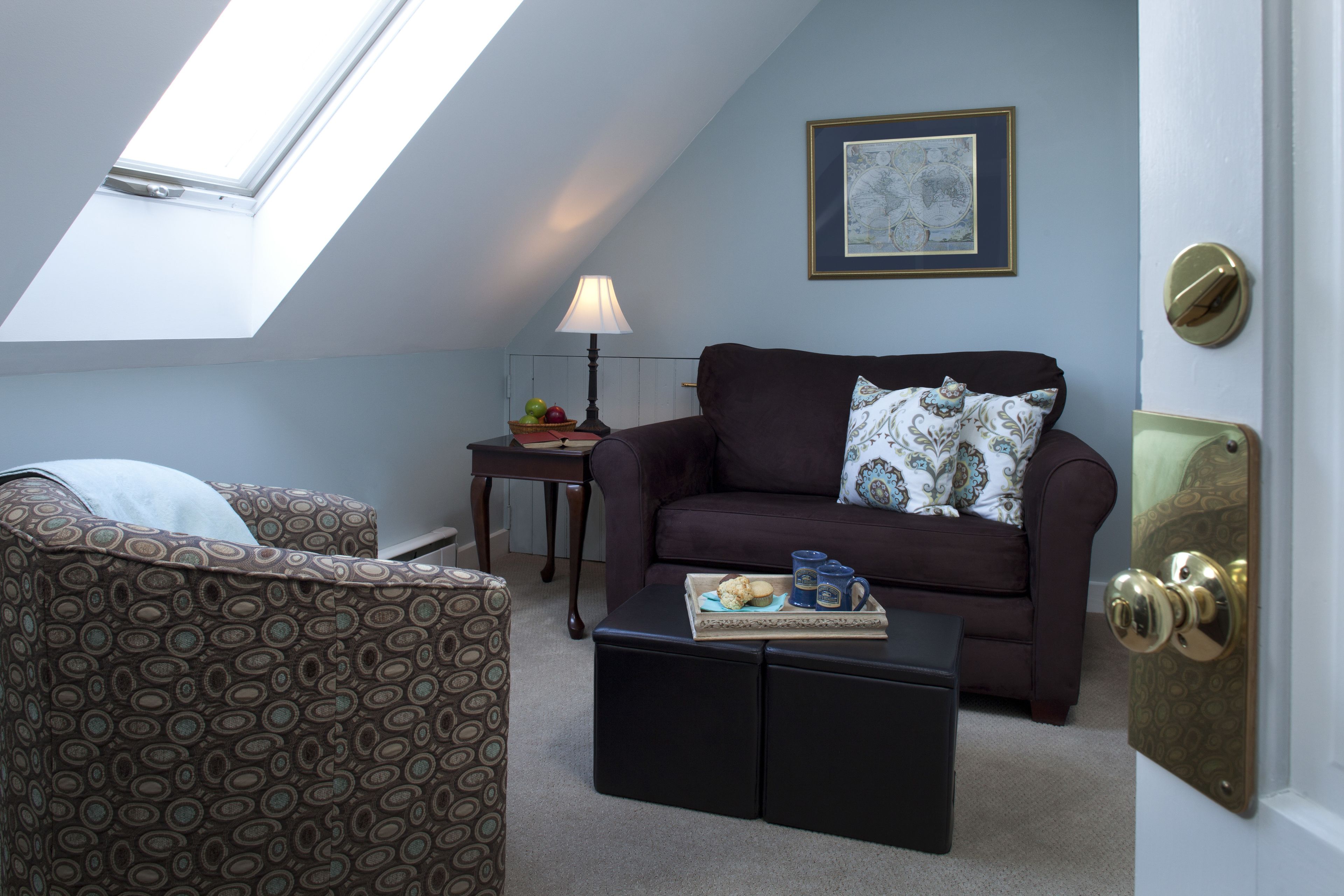 A cozy loft bedroom with a skylight, a dark brown sofa, a patterned armchair, and a small side table with a lamp and fruit.