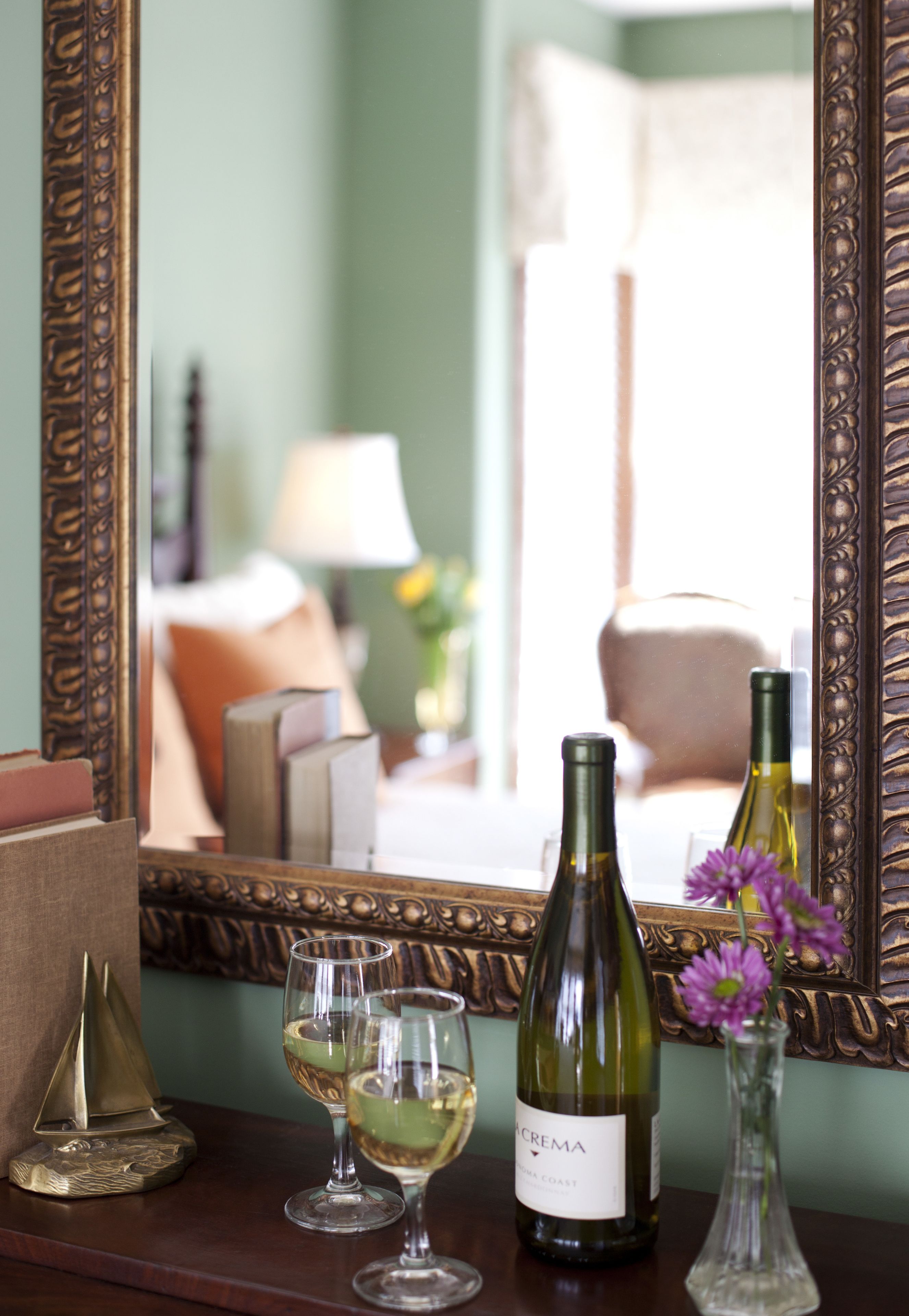 A mirror reflecting a room with a table holding a wine bottle, two glasses, and a vase with flowers.