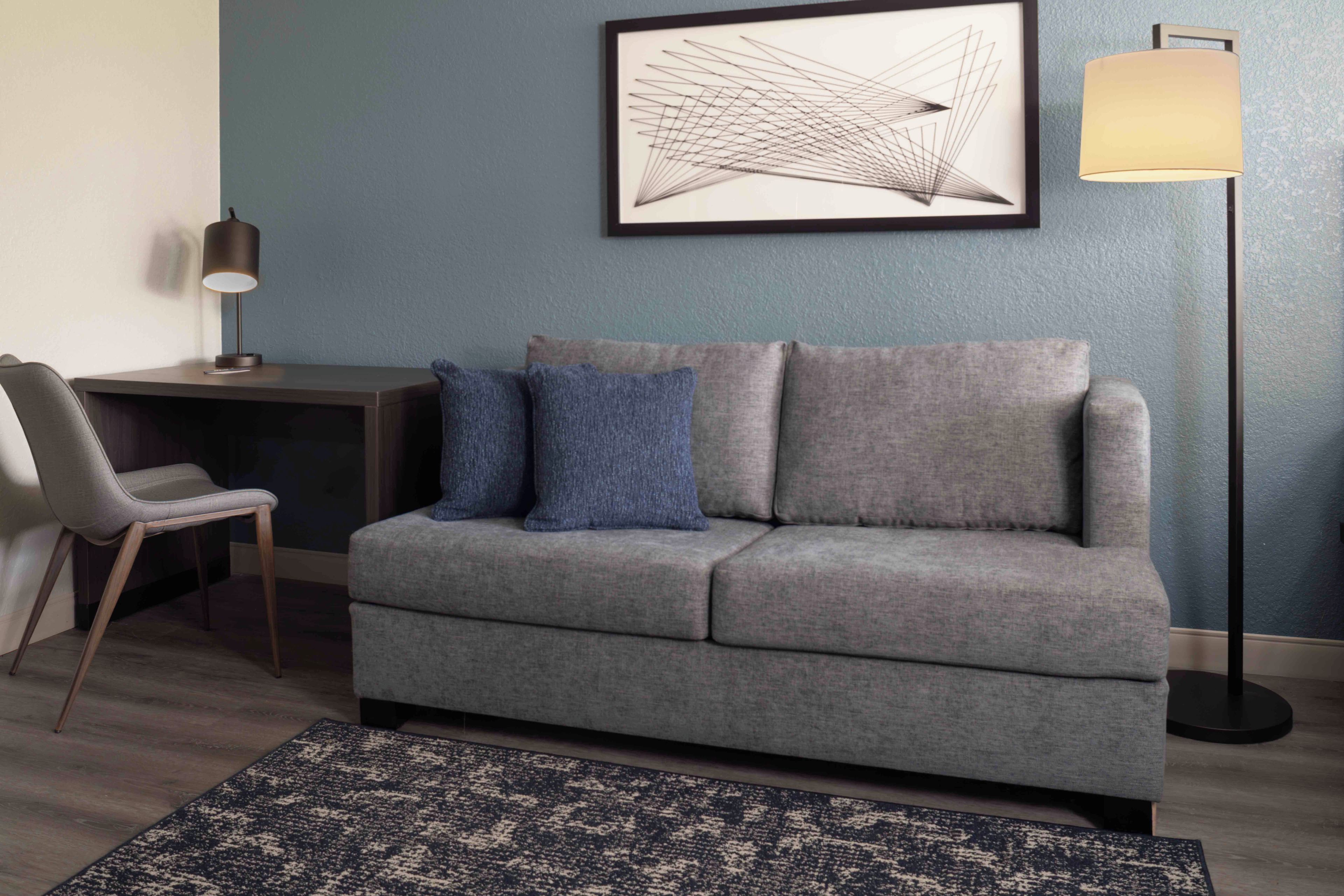 Una sala moderna con un sofá gris, una alfombra azul, un escritorio con una lámpara y una obra de arte abstracta enmarcada en la pared.
