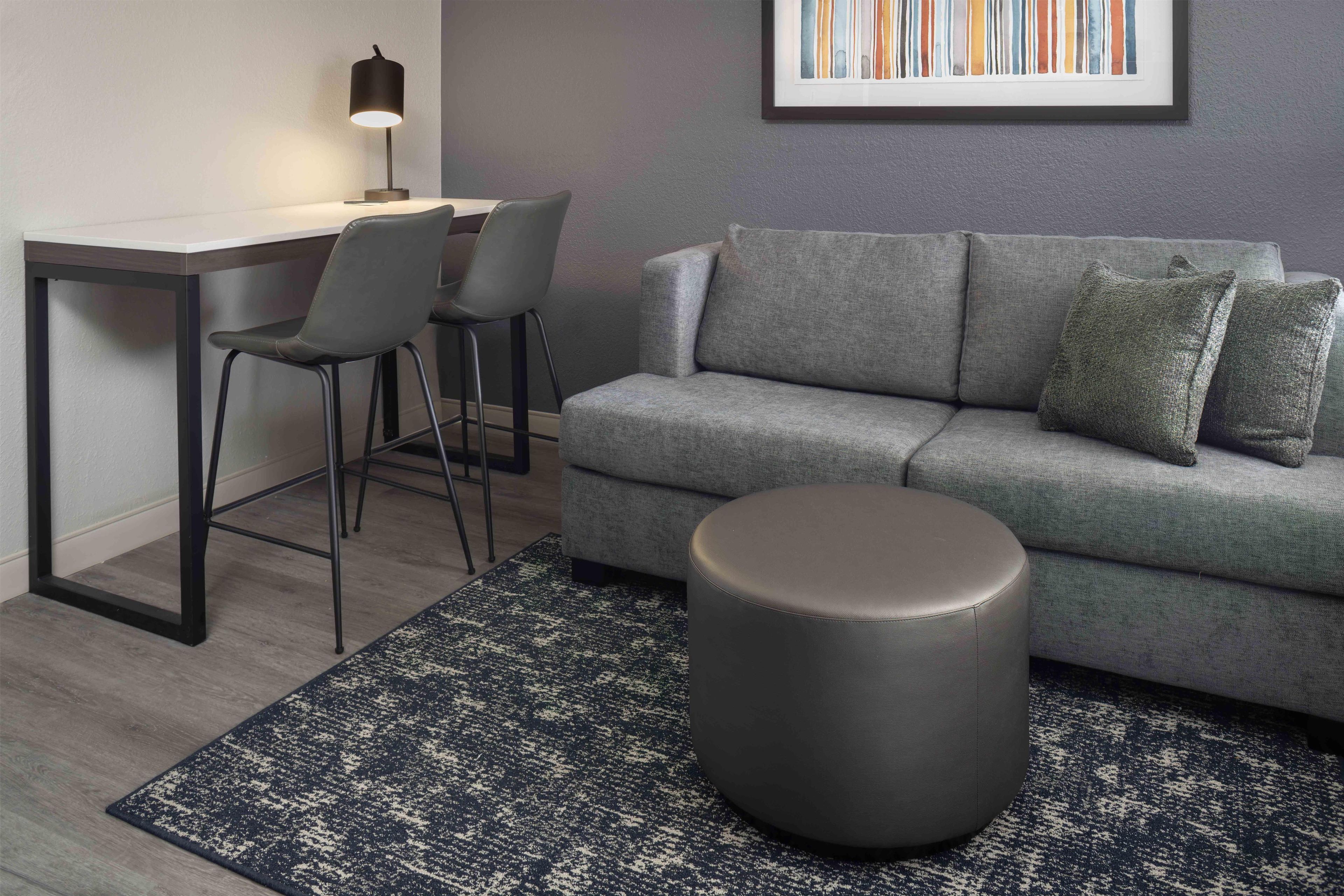 Un salon moderne avec un canapé gris, deux fauteuils, un pouf rond, un bureau avec une lampe et un tableau abstrait encadré accroché au mur.