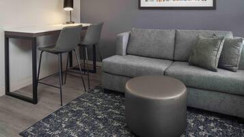 Un salon moderne avec un canapé gris, deux fauteuils, un pouf rond, un bureau avec une lampe et un tableau abstrait encadré accroché au mur.