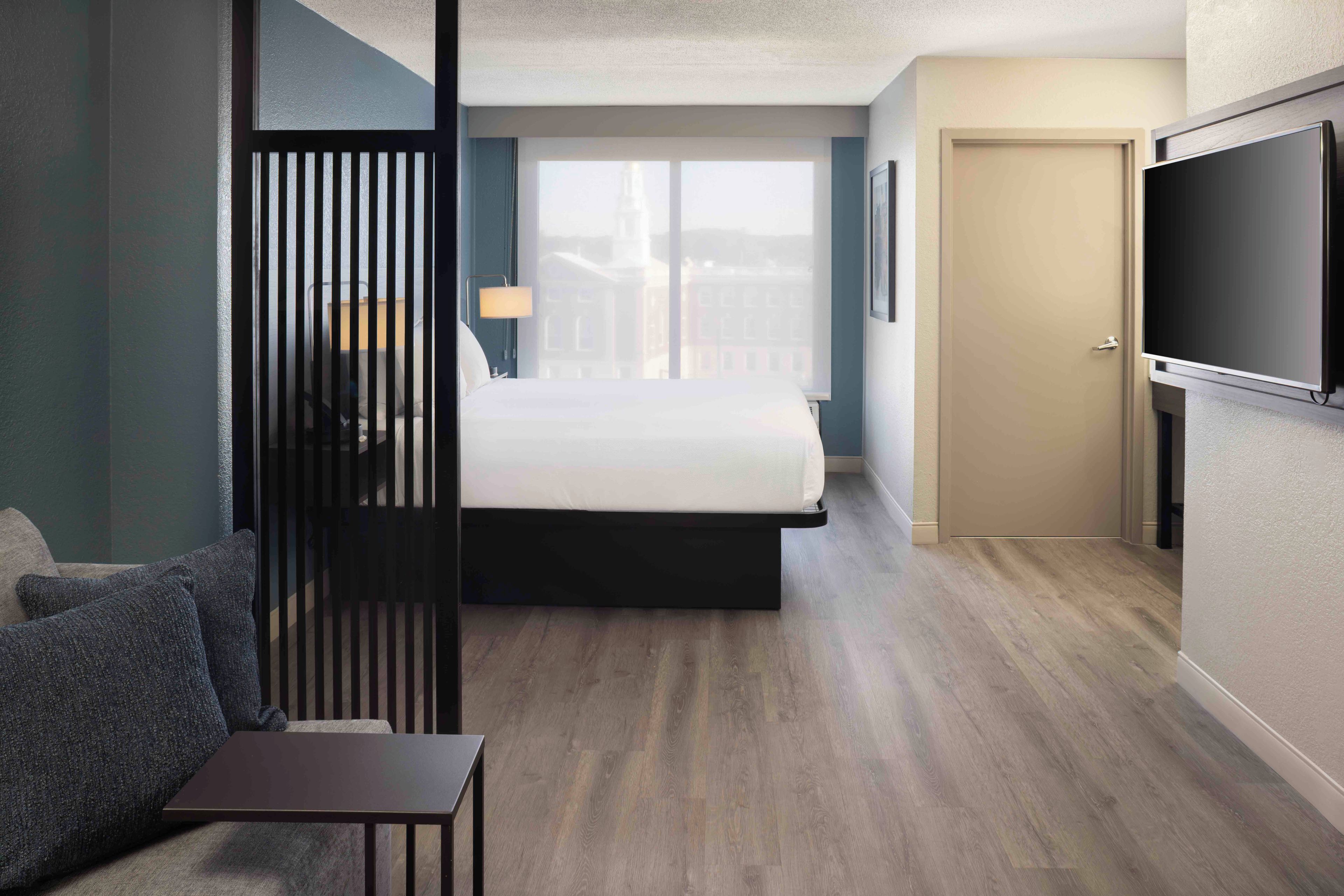 A hotel room with a bed, a TV, a desk, and a view of buildings through the window.