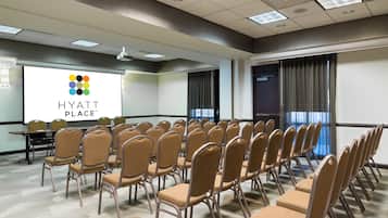 Un salón de conferencias con filas de sillas, una pantalla de proyección mostrando 'HYATT PLACE' y un amplio ventanal con cortinas.