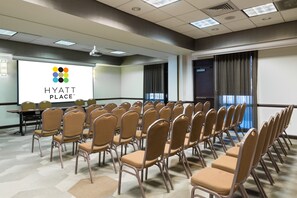 Una sala de conferències amb files de cadires, una pantalla de projecció que mostra 'HYATT PLACE' i un gran finestral amb cortines.