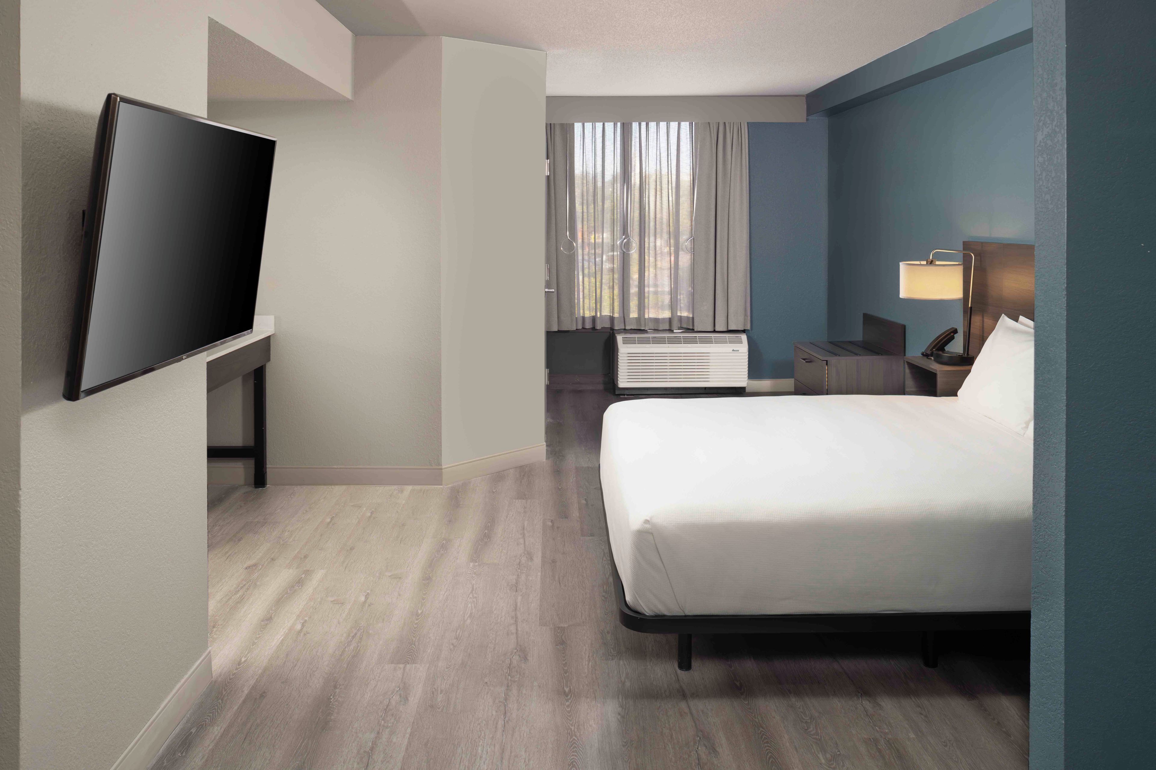 A hotel room with a bed, a flat-screen TV, a desk with a lamp, and a window with curtains.