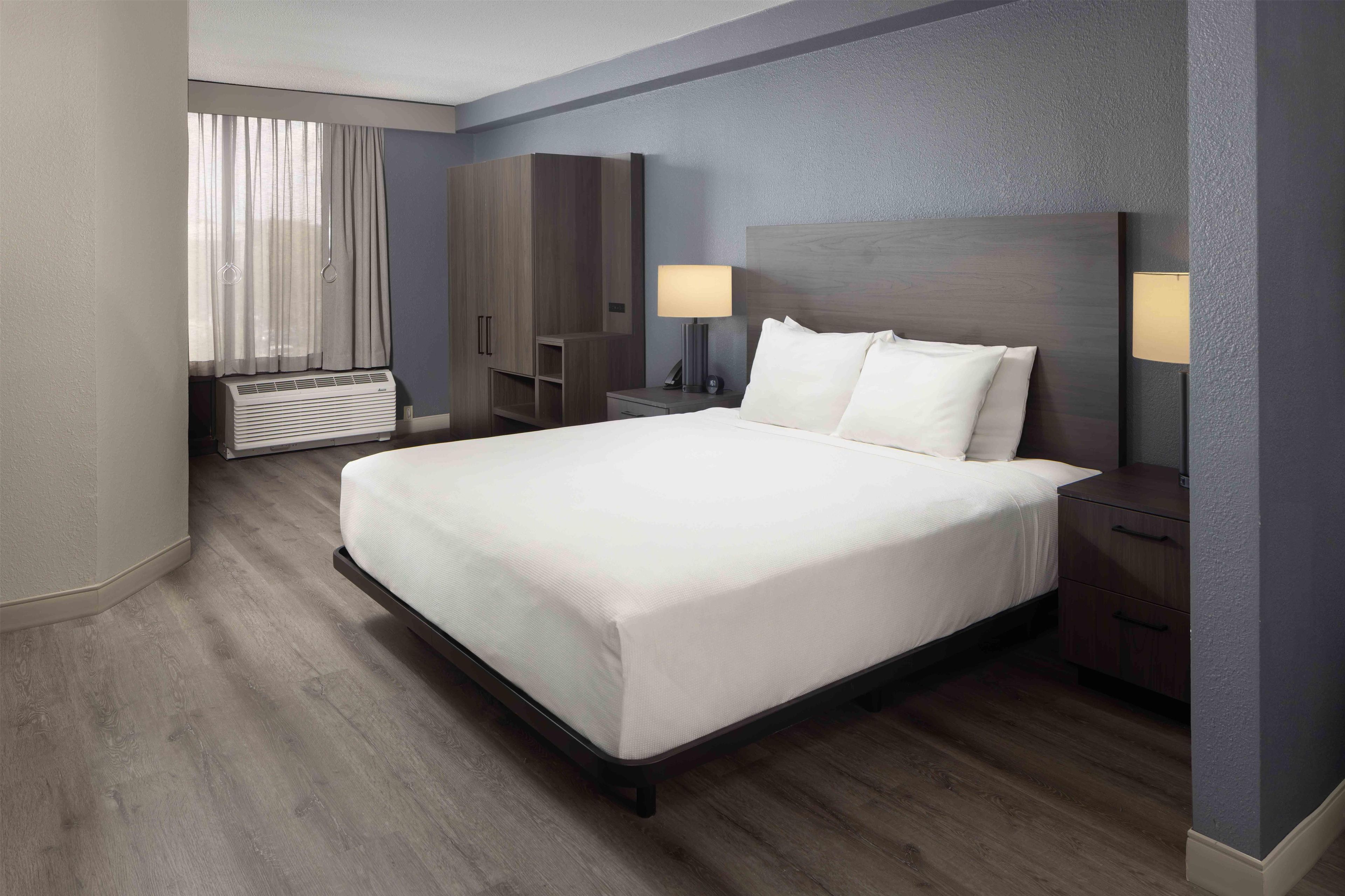 Una habitación de hotel con cama, lámparas de noche, armario y ventana con cortinas.