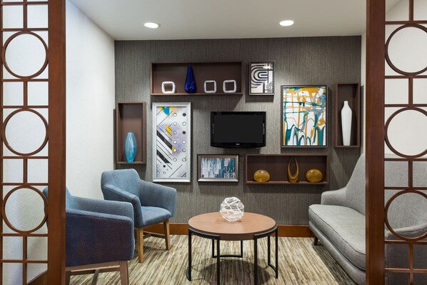A modern living room with a flat-screen TV, two blue armchairs, a grey sofa, and a round coffee table. - Hyatt Place Boston/Medford (Medford)