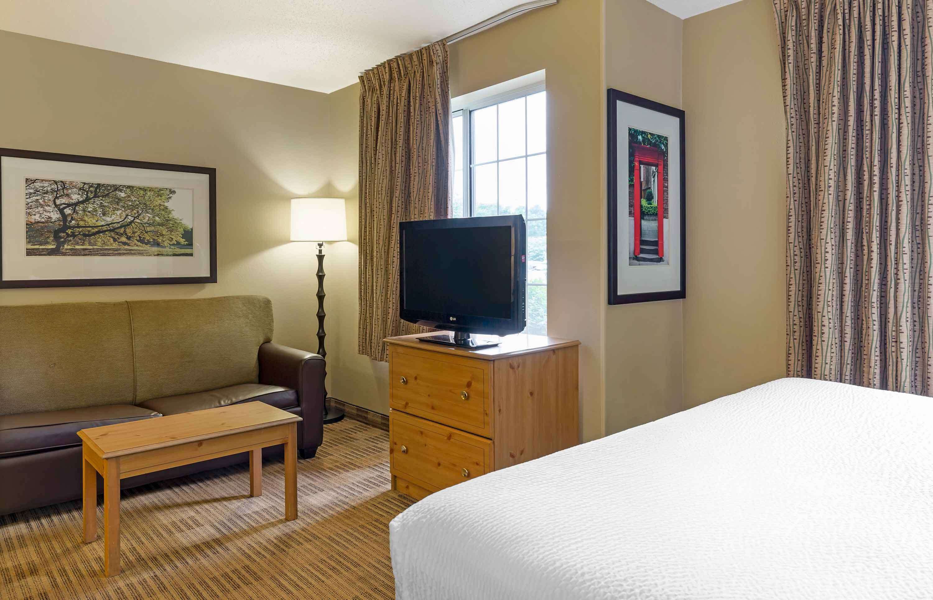 Habitación de hotel con cama, mesita de noche de madera, televisor, sofá, una pequeña mesa y ventana con cortinas.