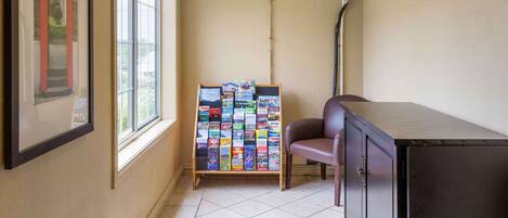 A hotel room with a large window, a wall-mounted air conditioner, a bulletin board with travel brochures, a leather chair, and a dark wooden cabinet.