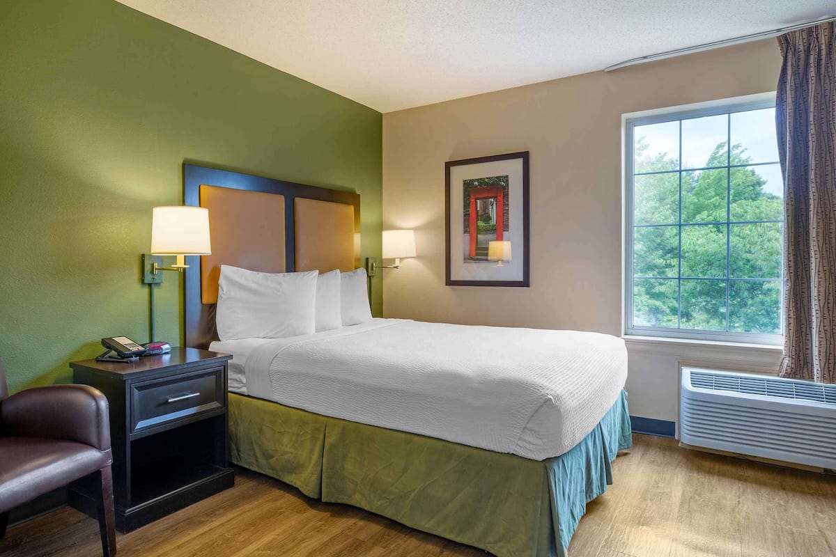 A hotel room with a bed, bedside lamps, a desk with a phone, a chair, and a window with a view of trees.