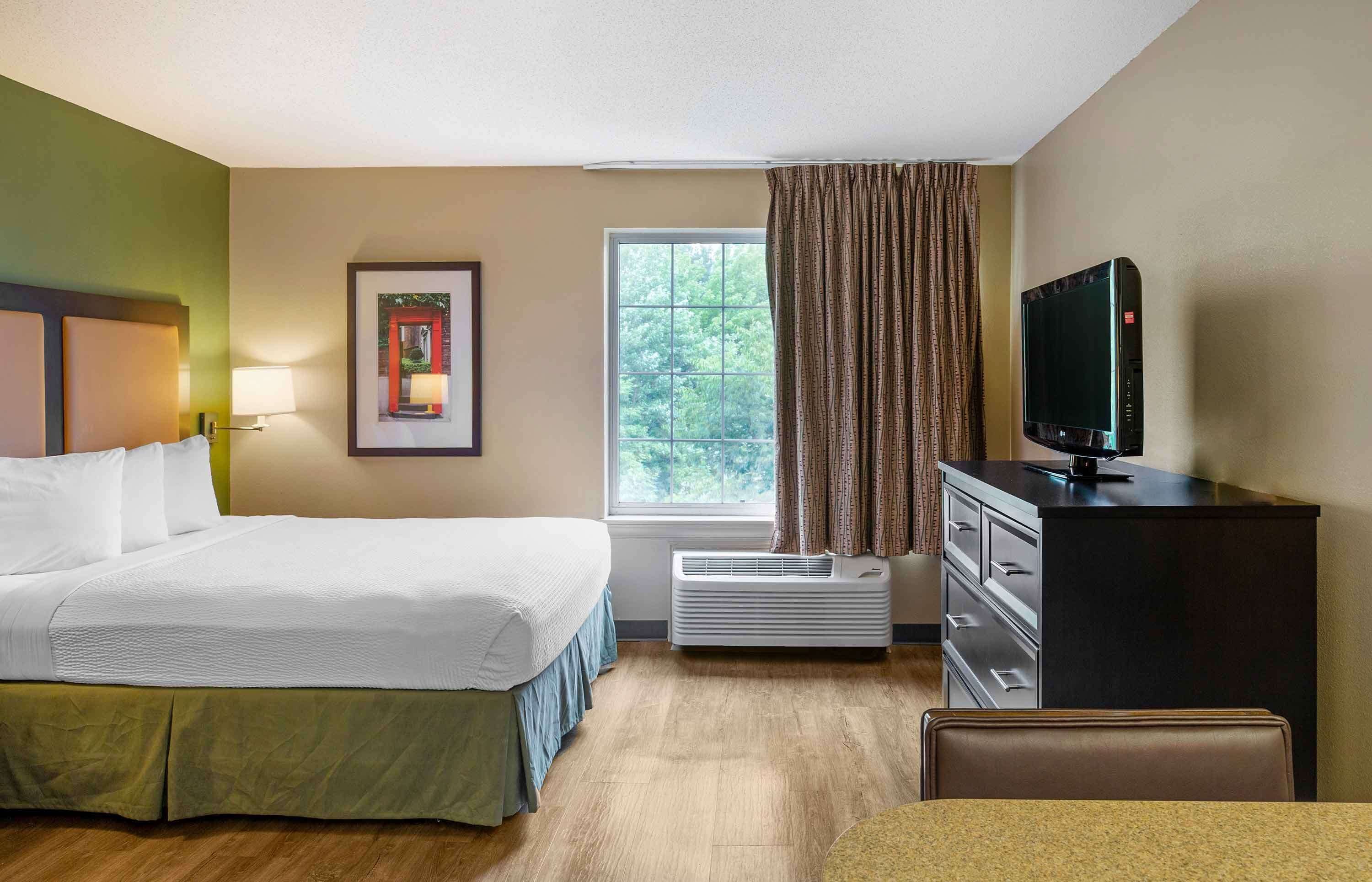 A hotel room with a bed, a desk with a television, a window with curtains, and a framed picture on the wall.