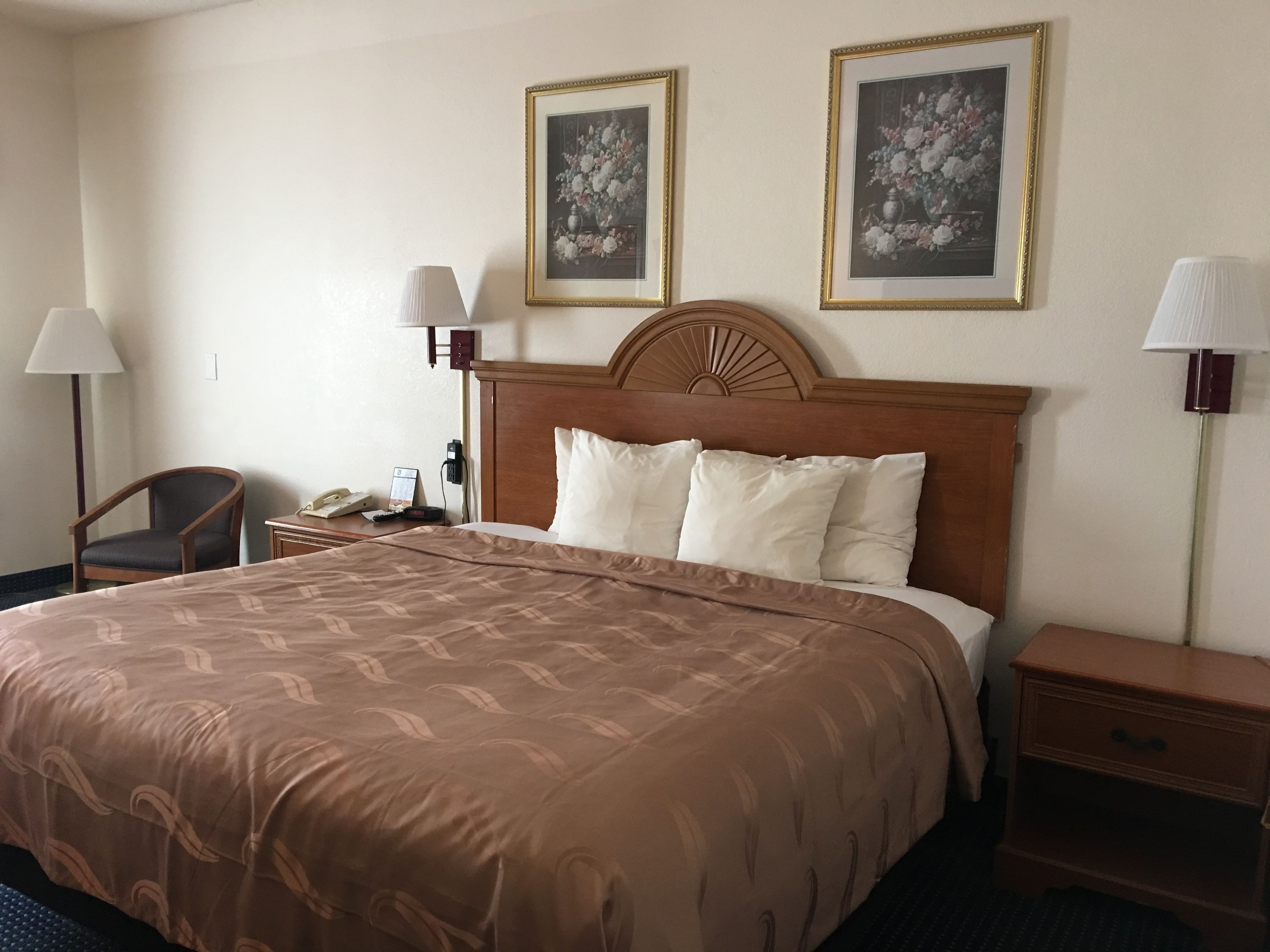 Habitación de hotel con una cama grande, dos lámparas de noche, una mesita de noche de madera, una silla y dos cuadros enmarcados con motivos florales en la pared.