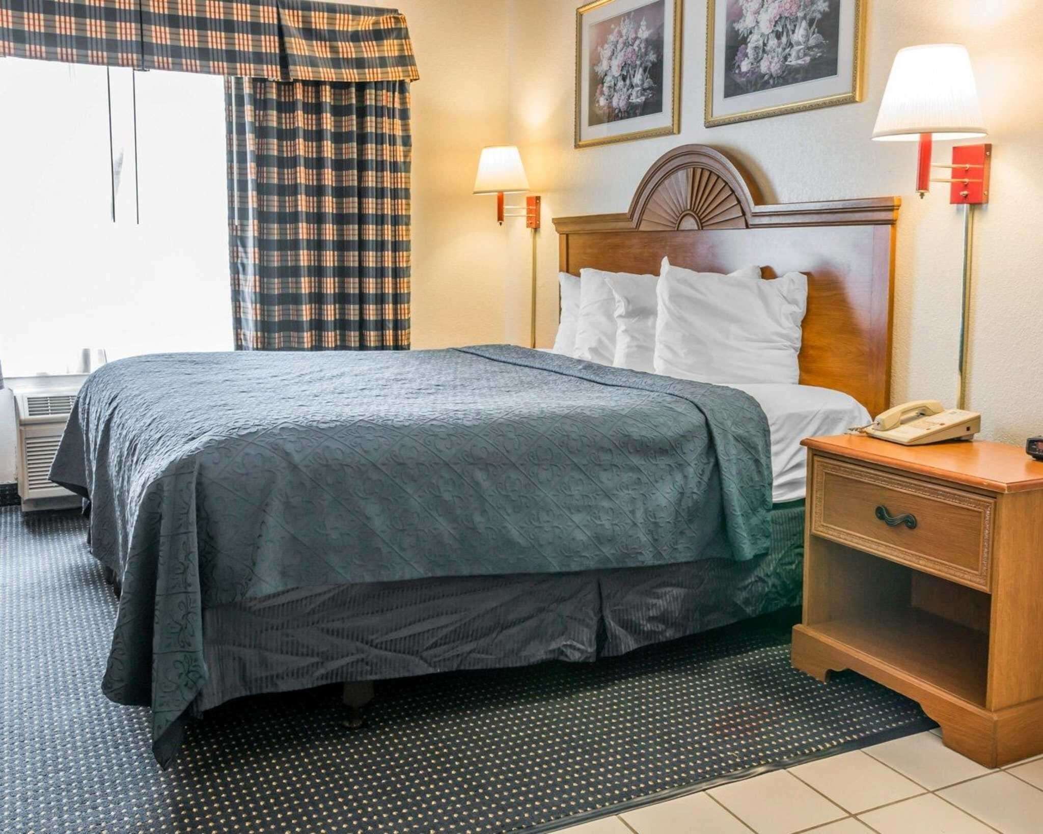 A hotel room with a bed, two bedside lamps, a nightstand with a phone, and a window with curtains.