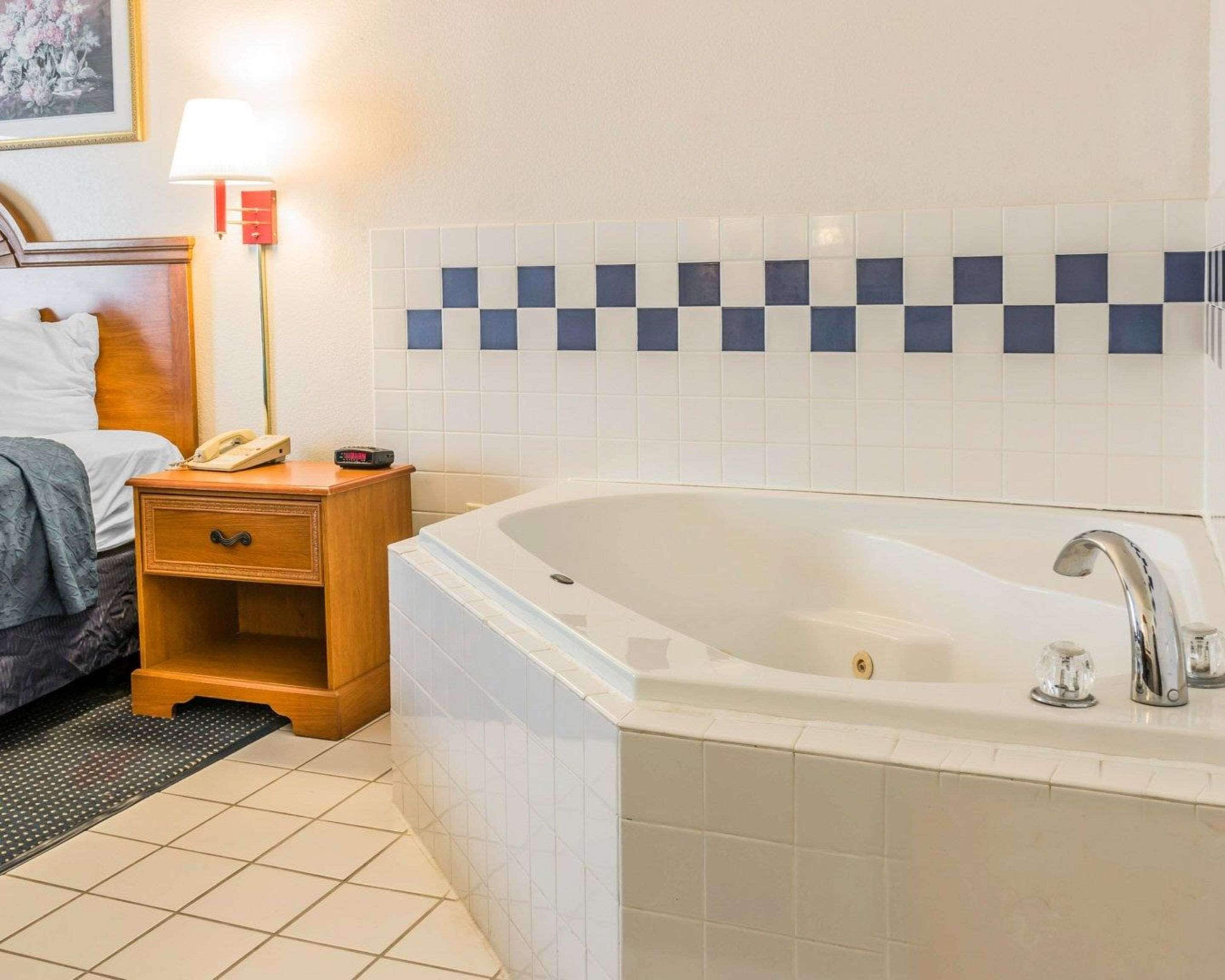 A hotel bathroom with a large bathtub, a bedside table with a lamp, and a tiled wall.