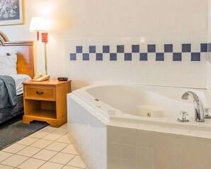 A hotel bathroom with a large bathtub, a bedside table with a lamp, and a tiled wall. - Quality Inn Kalamazoo near Wings Stadium (Kalamazoo)