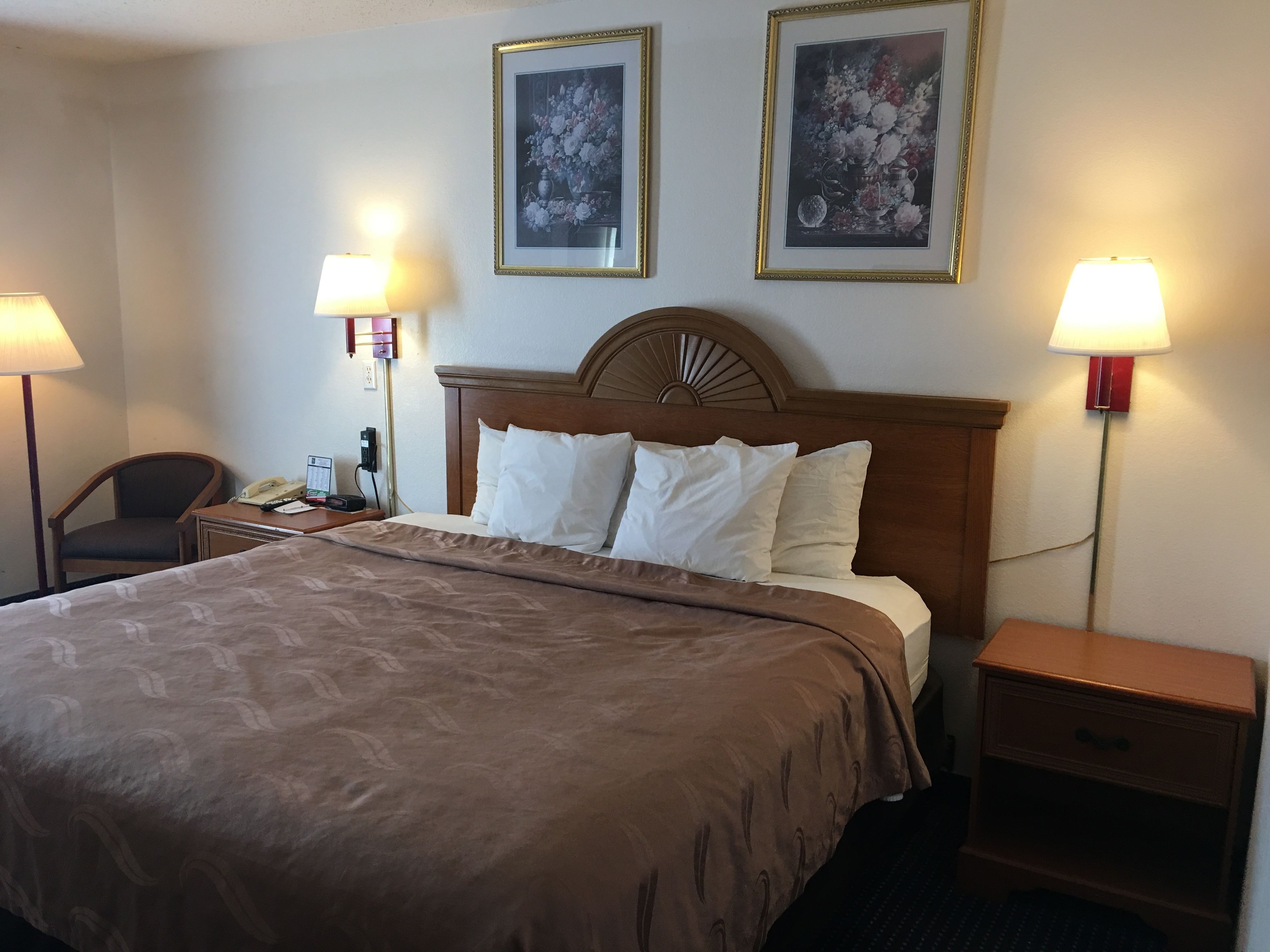 A hotel room with a large bed, two bedside lamps, a wooden headboard, and two framed floral paintings on the wall.