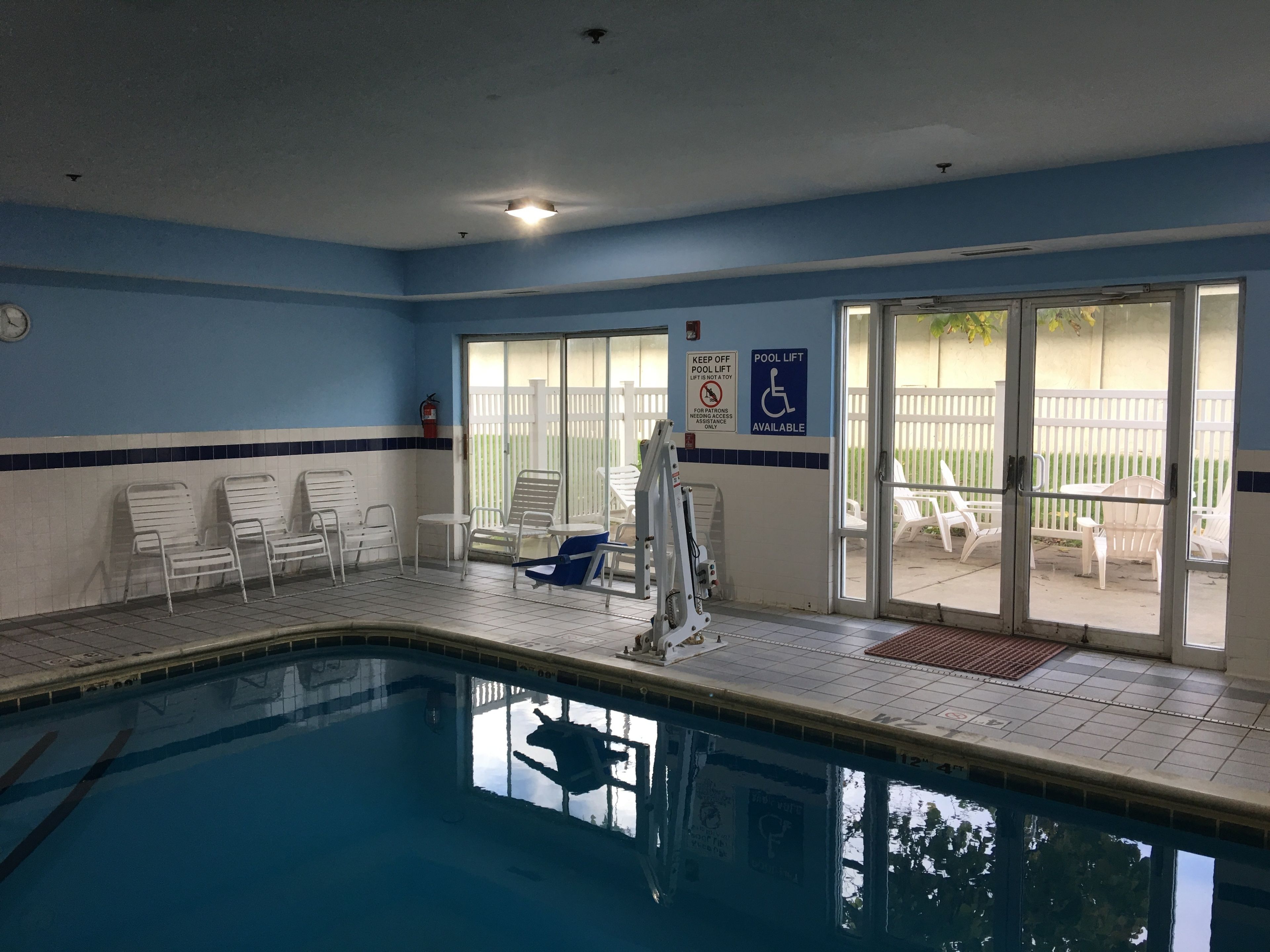 An indoor swimming pool with clear blue water, white lounge chairs, and a pool lift.