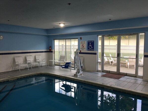 An indoor swimming pool with clear blue water, white lounge chairs, and a pool lift.