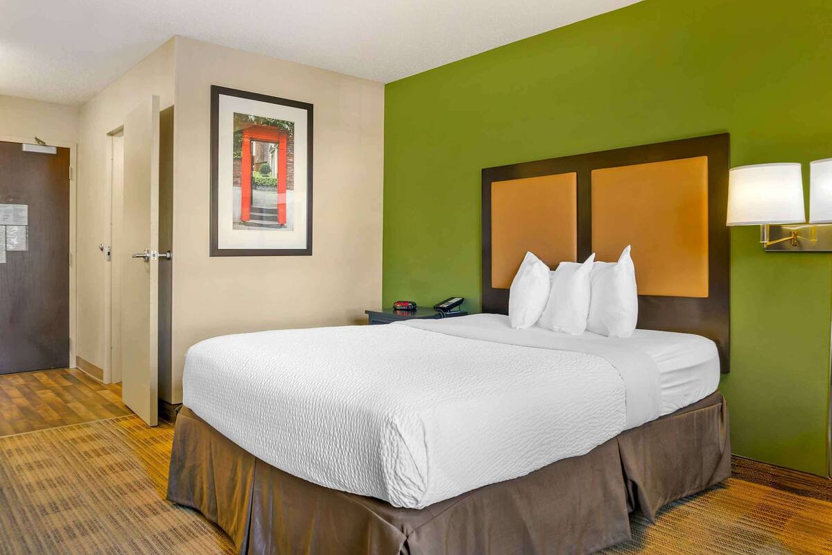 A hotel room with a large bed, a framed picture on the wall, a bedside table with a lamp, and a green accent wall.