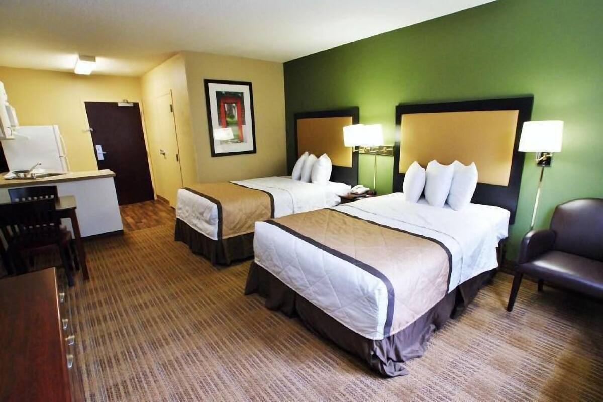 A hotel room with two beds, a desk, a chair, and a small kitchenette.