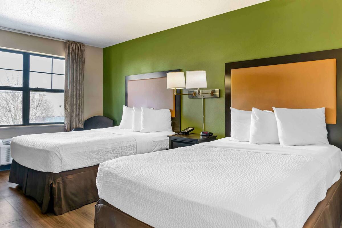 A hotel room with two beds, a window with curtains, a nightstand with a lamp, and a green wall.
