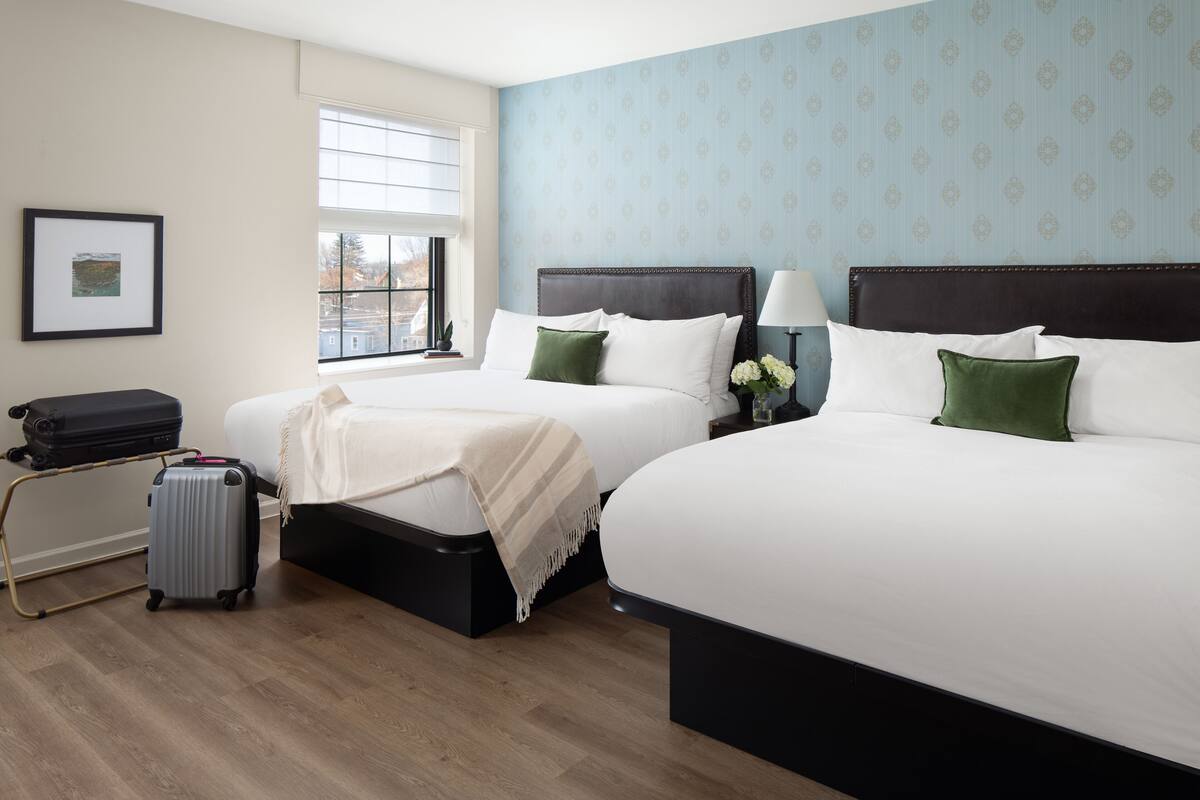 A hotel room with two beds, a window, a framed picture on the wall, and a suitcase on a trolley.
