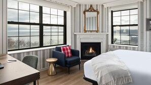 A hotel room with a large bed, a fireplace, a seating area with a chair and a small table, and large windows offering a view of the outdoors. - Landmark Inn (Marquette)